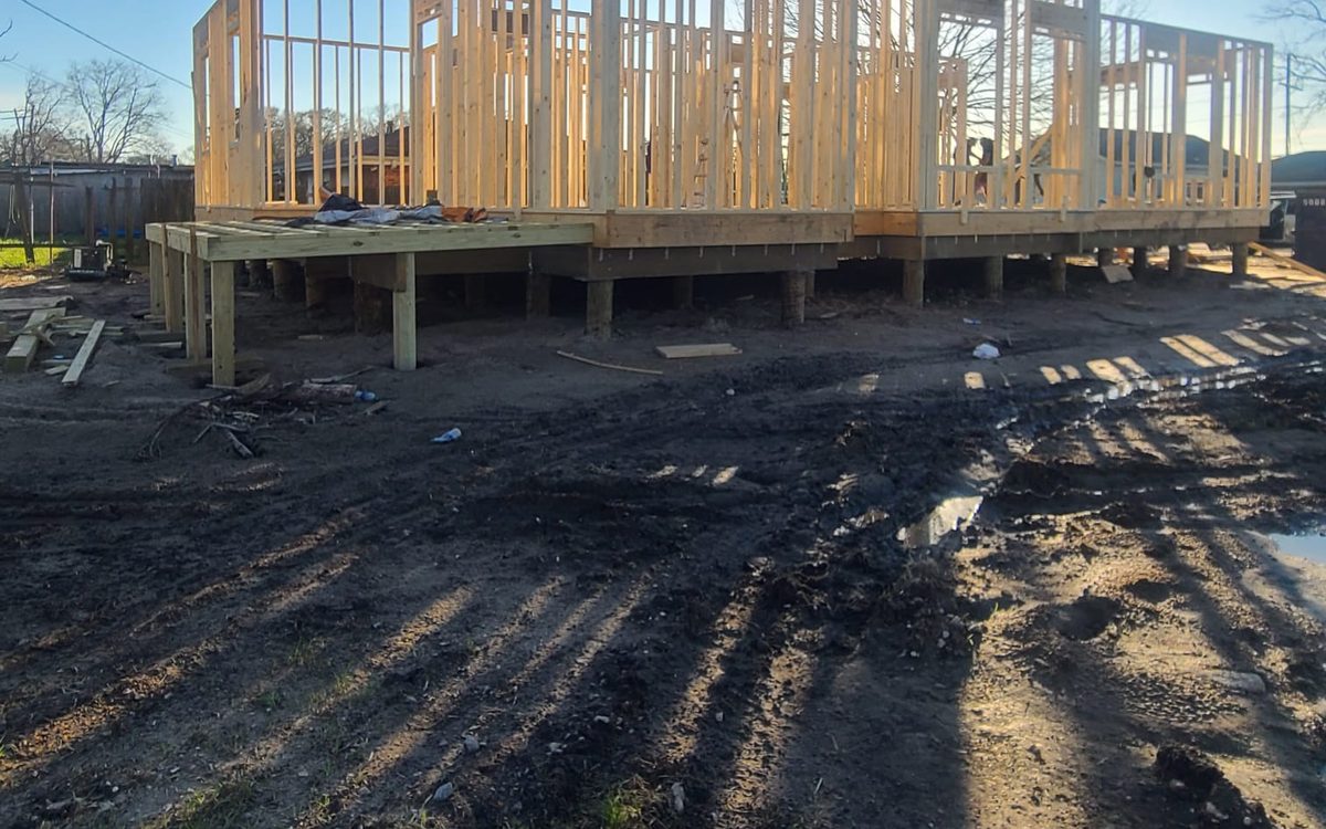 Wall framing on a raised platform supported by timber pilings, backlit by the sun