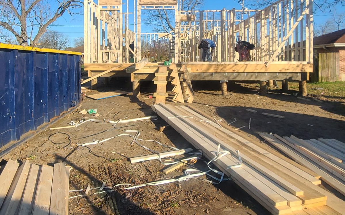Crew framing walls on a raised platform, with lumber stacked in the foreground