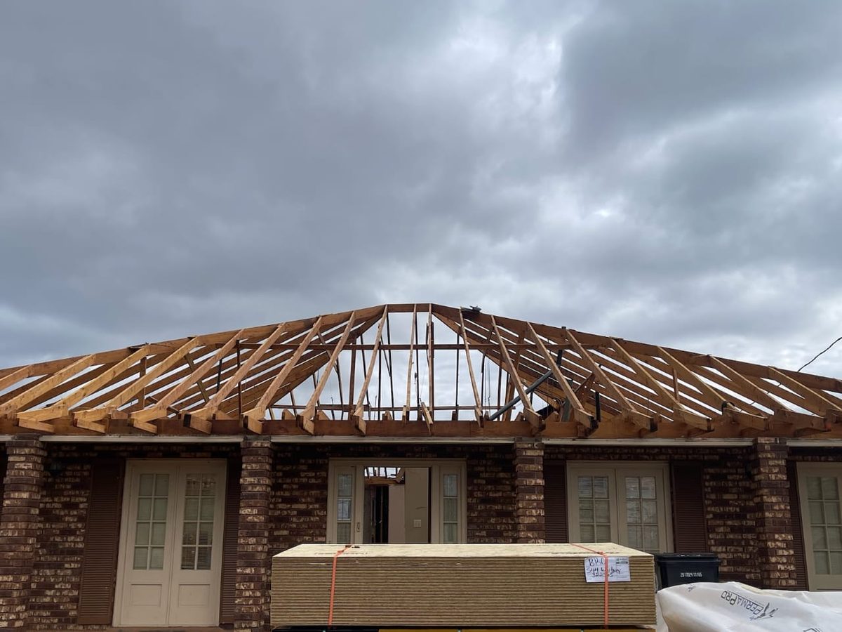 Exposed hip roof framing on a brick home, with subflooring panels staged in front