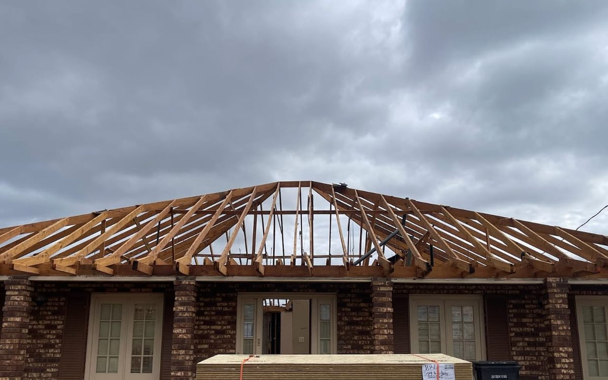 Exposed hip roof framing on a brick home, with subflooring panels staged in front